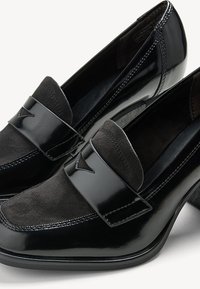 Mocassins en cuir verni noir et en daim avec des talons bas et des brides décoratives sur le devant, vus de près sous un angle.