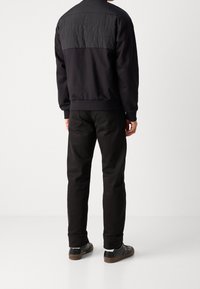 Chaqueta bomber negra con espalda texturizada, puños y dobladillo acanalados, combinada con jeans negros y zapatos negros, vista desde la espalda.