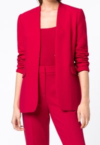 Blazer rosso realizzato in tessuto strutturato, con rever a punta, apertura frontale e tasche frontali. Abbinato a un top e pantaloni coordinati.