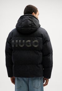 Svart puffjakke med stor hette og "HUGO"-logo på baksiden. Har et strukturert materiale og en firkantet silhuett. Løs stil.