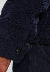 Granatowa polarowa kurtka z teksturowaną powierzchnią, z czarnym guzikiem na mankiecie oraz gładkim materiałem pod spodem.