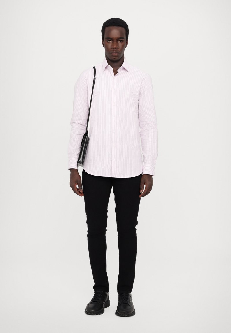 Hellrosa gestreiftes Hemd mit Knöpfen, schlanke schwarze Hose, schwarze Schuhe und eine kleine schwarze Umhängetasche. Das Modell steht vor einem weißen Hintergrund.