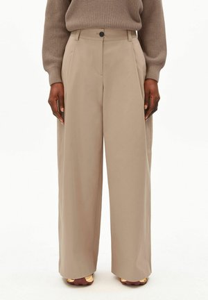 Modell trägt beige weite Hosen, kombiniert mit einem braunen Strickpullover und beigen Turnschuhen, stehend vor einem schlichten Hintergrund.