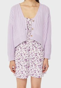 Cardigan en maille côtelée de couleur violet clair avec trois boutons, superposé sur une robe blanche ornée d'un petit motif floral violet. Manches courtes visibles.