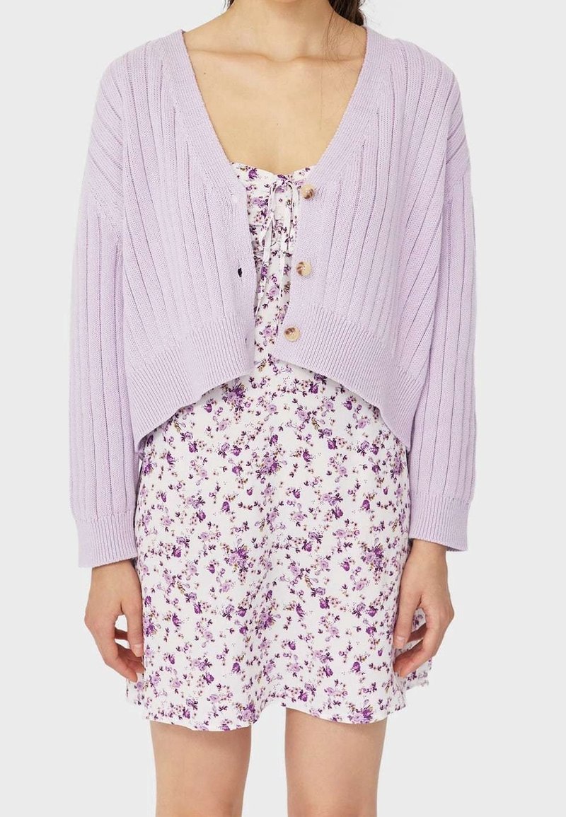 Cardigan en maille côtelée de couleur violet clair avec trois boutons, superposé sur une robe blanche ornée d'un petit motif floral violet. Manches courtes visibles.
