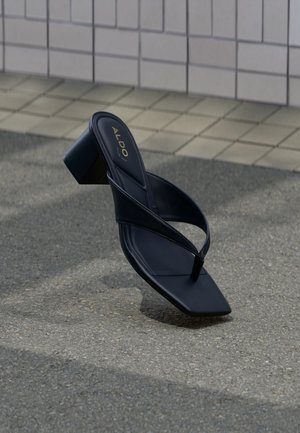 Sandale negre cu toc, cu vârf pătrat și barete late, expuse pe un perete placat cu faianță și o suprafață texturată a solului.