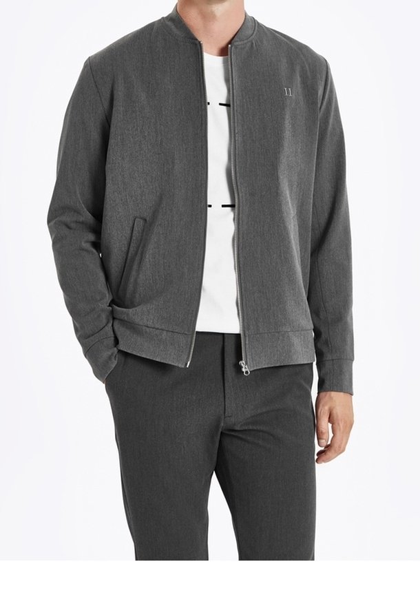 Homme portant une veste grise zippée par-dessus une chemise blanche et un pantalon gris, debout avec une main dans la poche et l'autre détendue.