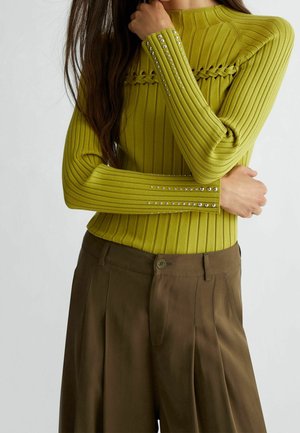 Žena v žebrovaném limetkově zeleném svetru s cvočky na manžetách a pleteným detailem, doplněném o volné hnědé kalhoty s vysokým pasem, se zkříženýma rukama.