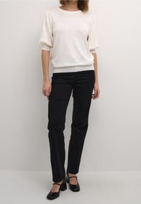 Suéter blanco de punto con mangas abullonadas, combinado con jeans negros y zapatos de tacón negros. Diseño sencillo con detalles en canalé en los puños y el dobladillo.
