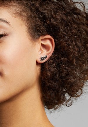 Oreille avec trois petites boucles d'oreilles noires en forme d'étoile sur une personne aux cheveux bruns bouclés et à la peau brun clair.