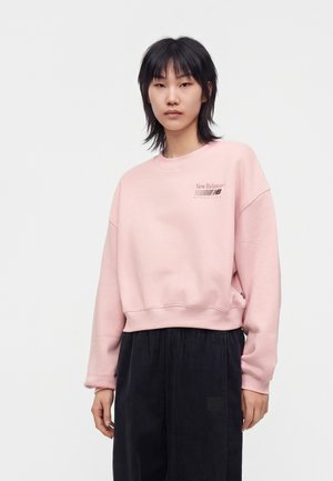 Ung person med sort hår iført lyserød New Balance sweatshirt og løse sorte bukser, stående mod en ensfarvet lys baggrund.