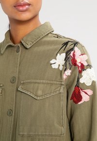 Olivově zelená bunda s texturovanou tkaninou, zdobená květinovou výšivkou v růžové a bílé barvě na levém rameni a dvěma předními kapsami.