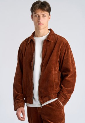 Young man wearing brown corduroy jacket and matching pants over a white t-shirt, standing against plain light background.