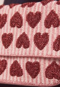 Gyöngyözött kézitáska rózsaszín szövetalappal, kinagyított piros szívmintákkal, váltakozó csíkos texturált háttér előtt.