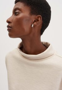 Maglione beige morbido a collo alto con una finitura testurizzata, caratterizzato da un colletto aderente e sottili dettagli a coste. I orecchini sono in argento lucido.
