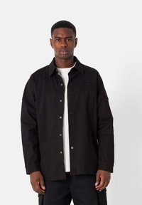 Black button-up jacket in a relaxed fit, featuring a collar, chest pocket, and snap buttons on the front. Worn over a white shirt.