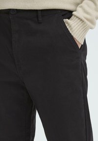 Black cotton trousers with a straight cut; features a button closure, pockets, and a soft texture. Ribbed beige sweater visible at the cuff.