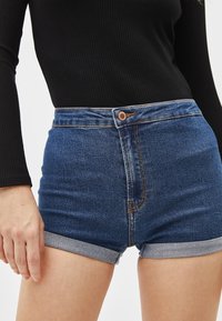 Shorts in denim a vita alta, indossati con l'orlo ripiegato, dotati di chiusura frontale con bottone e cuciture a contrasto in blu scuro.