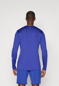 Ilga rankovė sportinė marškinė ryškiai mėlyna spalva, su lygiu paviršiumi. Yra apskritas akcentas nugaros dalyje ir logotipo detalė ant rankovės.