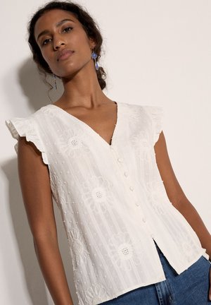 Mujer con blusa blanca sin mangas con bordado floral y pendientes azules colgantes, de pie contra un fondo claro.