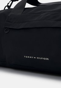 Borsa a tracolla nera in nylon con una superficie liscia, dotata di manico superiore, tasca laterale e logo bianco "TOMMY HILFIGER" sul davanti.