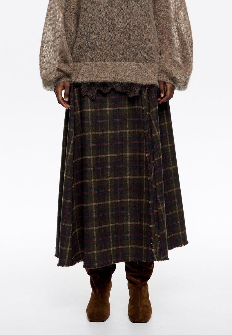 Sukňa strednej dĺžky s zeleno-námorníckym károvaným vzorom, skombinovaná s hnedými semišovými čižmami a voľným hnedým pleteným sveterom.