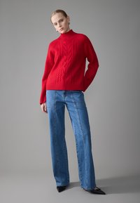 Roter Strickpullover mit erhabenem Zopfmuster, hochgeschlossener Kragen, kombiniert mit hellblauen weit geschnittenen Jeans und schwarzen spitzen Schuhen.