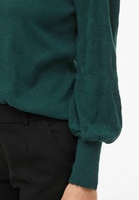 Maglione lavorato a maglia in teal con una finitura strutturata e maniche a sbuffo, abbinato a pantaloni neri, che mette in risalto la morbidezza e la forma del capo.