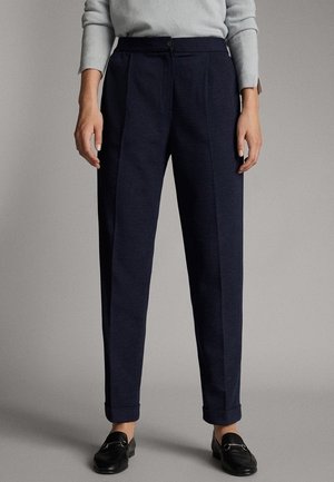 Personne portant un pantalon plissé bleu marine, un pull gris clair à manches longues et des mocassins noirs, debout devant un fond neutre.