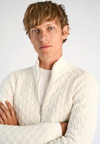 Vit zip-up tröja med ett strukturerat diamantmönster, med ribbad krage och manschetter, som bärs över en vit t-shirt.