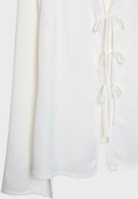 Vêtement blanc à manches longues avec des fentes latérales nouées par plusieurs petits nœuds le long de la couture.