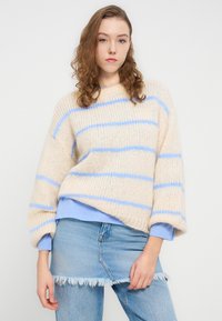 Πλεκτό πουλόβερ σε κρεμ απόχρωση με μπλε ρίγες, με φαρδιά εφαρμογή και φαρδιά μανίκια. Φορεμένο πάνω από ένα ανοιχτό μπλε πουκάμισο, σε συνδυασμό με μια φούστα από τζιν με φθαρμένα τελειώματα.