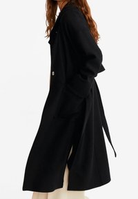 Černý dlouhý kabát vyrobený z texturované látky. Má šálový límec, dvě přední kapsy a opasek na zavázání v pase.