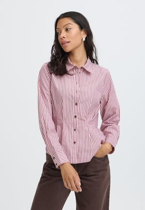 Femme aux cheveux foncés porte une chemise à rayures verticales roses et noires boutonnée et un pantalon marron foncé, posant avec une main dans la poche.