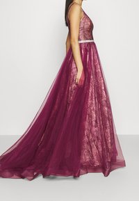 Długa wieczorowa sukienka w ciemnym burgundzie, z dopasowanym koronkowym stanikiem, przezroczystą tiulową spódnicą i delikatnym detalem w pasie.