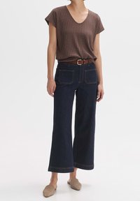 Hnědé tričko s krátkými rukávy a vzorem, s výstřihem do V, kombinované s tmavě modrými džínami s širokými nohavicemi a hnědým opaskem. Měkká textura, pohodlný střih.
