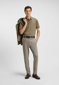 Khaki poloshirt met kraag en subtiel logo, gecombineerd met lichtgrijze op maat gemaakte broek en bruine leren schoenen. Casual jas over de schouder gedrapeerd.