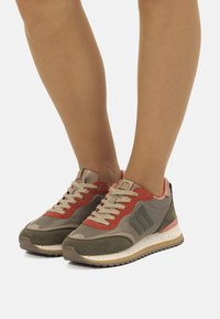 Sportliche Sneakers mit einer Mischung aus olivgrünem, rotem und beigem Material, strukturiertem Stoff, Gummisohle und Schnürdesign mit Logo-Akzenten.
