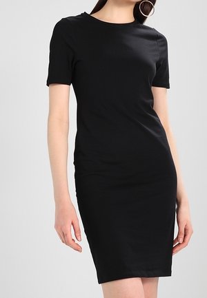 Femme portant une robe noire ajustée, à manches courtes, longueur genou, avec un col rond et de grandes boucles d'oreilles, sur un fond uni.