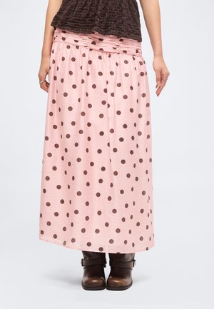 Nainen, joka pukeutunut vaaleanpunaiseen maksihameeseen, jossa on ruskeita pilkkuja, ruskeisiin nilkkasaappaisiin ja tummanruskeaan teksturoituun yläosaan, seisoo käsivarret rentoina.