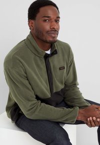 Green fleece pullover with a black lower panel and a half-zip closure. Features a high collar and a brand label on the left chest area.