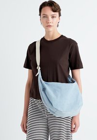 Hellblaue, lässige Denim-Tasche mit einem cremefarbenen Schultergurt; hat eine runde Form und dezente Nähte; wird crossbody getragen.