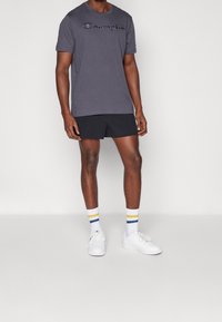 Camiseta gris de manga corta con un logo negro de "Champion", combinada con pantalones cortos negros, zapatillas blancas y calcetines rayados en tres colores.