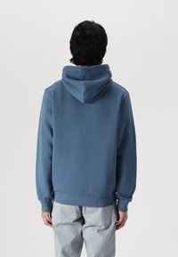 Felpa blu con vestibilità rilassata, ampio cappuccio, polsini e orlo a coste, realizzata in misto cotone. Indossata con pantaloni grigi chiari.