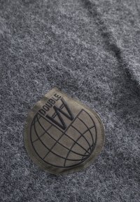Grå filtstof med et struktureret patch med et globusdesign og "DOUBLE W" tekst i mørkebrunt, syet langs kanten.