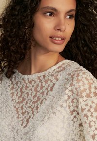 Haut en dentelle blanche avec un col rond et un tissu transparent. De petites touches jaunes et un motif texturé sont visibles sur l'ensemble.