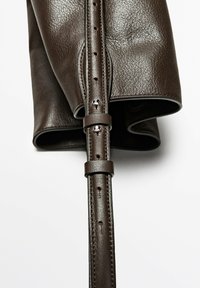 Bandoulière de sac à main en cuir texturé marron foncé avec clous argentés et bords cousus sur fond blanc.