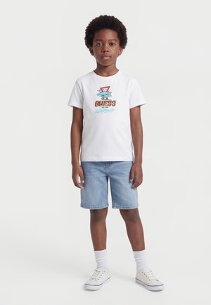 Αγόρι που φοράει λευκό T-shirt με κοντά μανίκια, πολύχρωμο γραφικό και κείμενο, ανοιχτό μπλε τζιν σορτς, λευκές κάλτσες και λευκά αθλητικά παπούτσια, όρθιο.