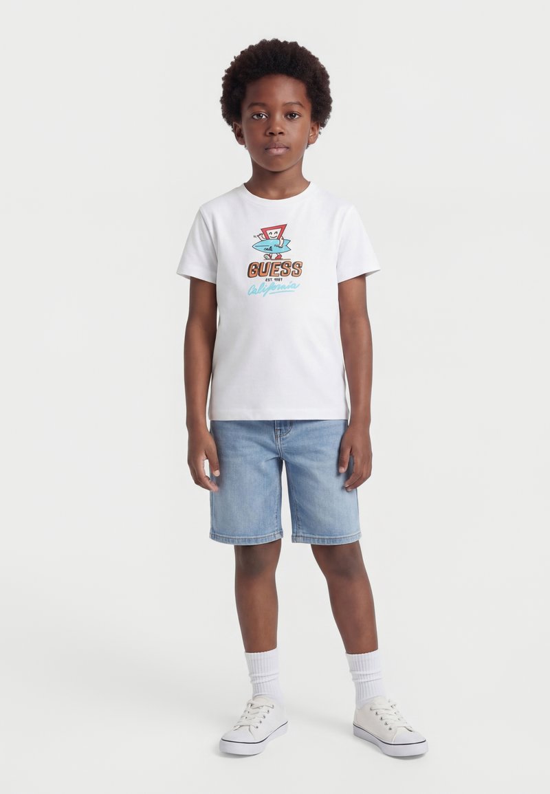 Αγόρι που φοράει λευκό T-shirt με κοντά μανίκια, πολύχρωμο γραφικό και κείμενο, ανοιχτό μπλε τζιν σορτς, λευκές κάλτσες και λευκά αθλητικά παπούτσια, όρθιο.