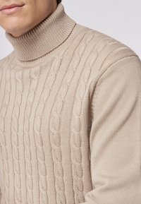 Beige Kabelstrick-Pullover mit Rollkragen, der strukturierte Muster und glatte Strickabschnitte für einen Schichteffekt aufweist.